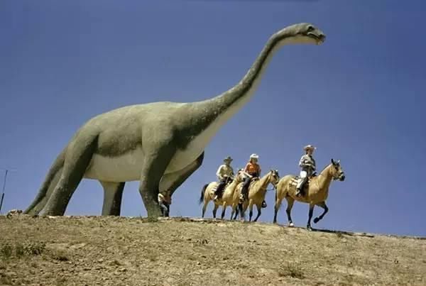 1956年,美国,骑着马的游客经过南达科他州恐龙园内一只逼真的恐龙模型