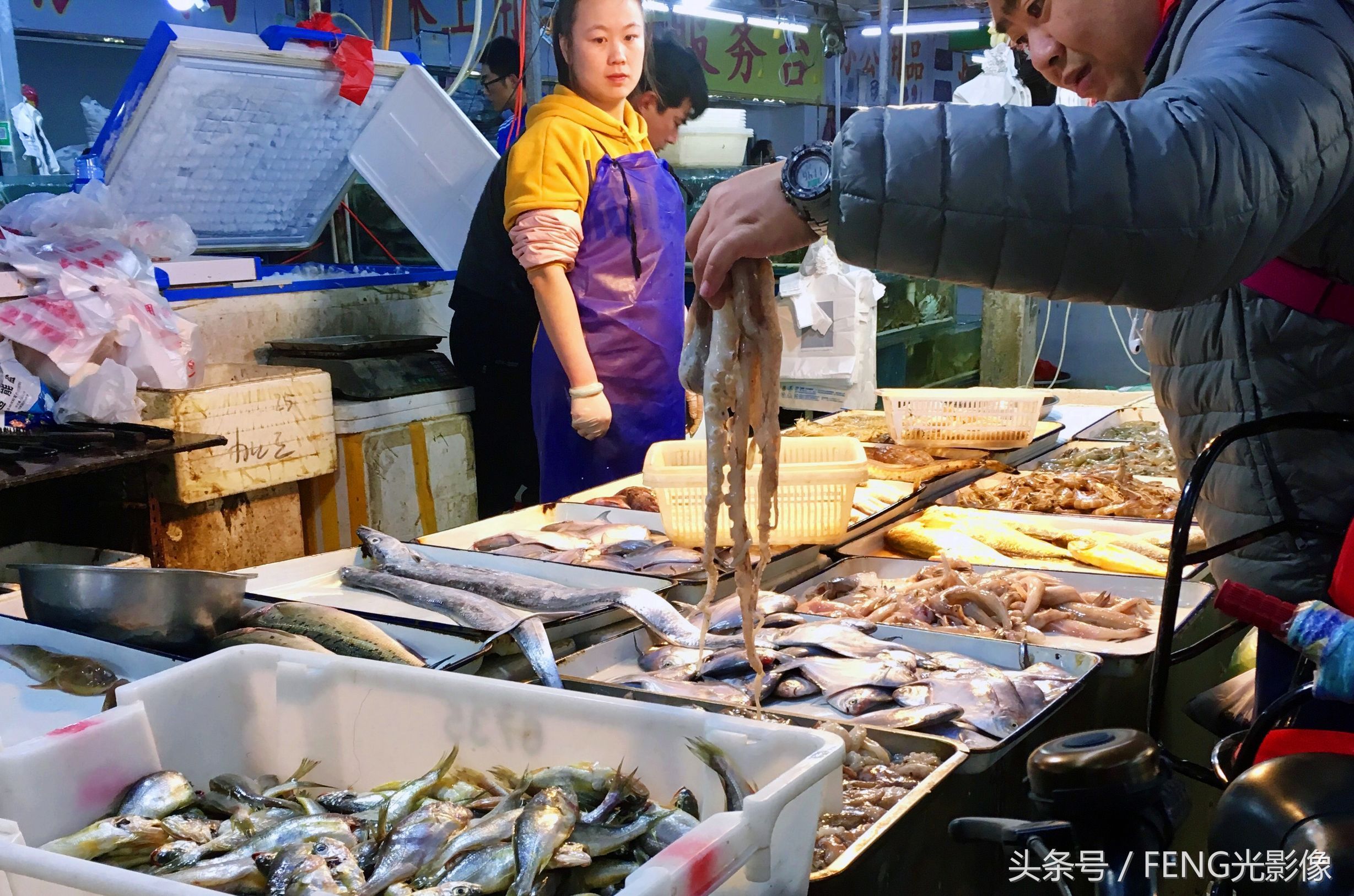 一位顾客在海鲜摊挑选八抓鱼,老板娘在一旁观望,等候为其称重结账